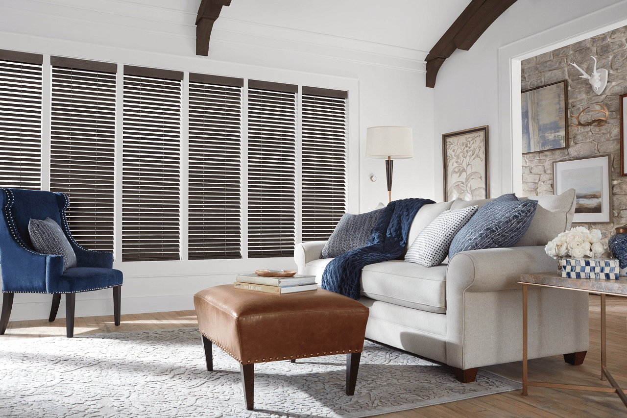 Stylish living room with dark wood blinds covering a row of windows, paired with a beige sofa, blue accent chair, and leather ottoman.
