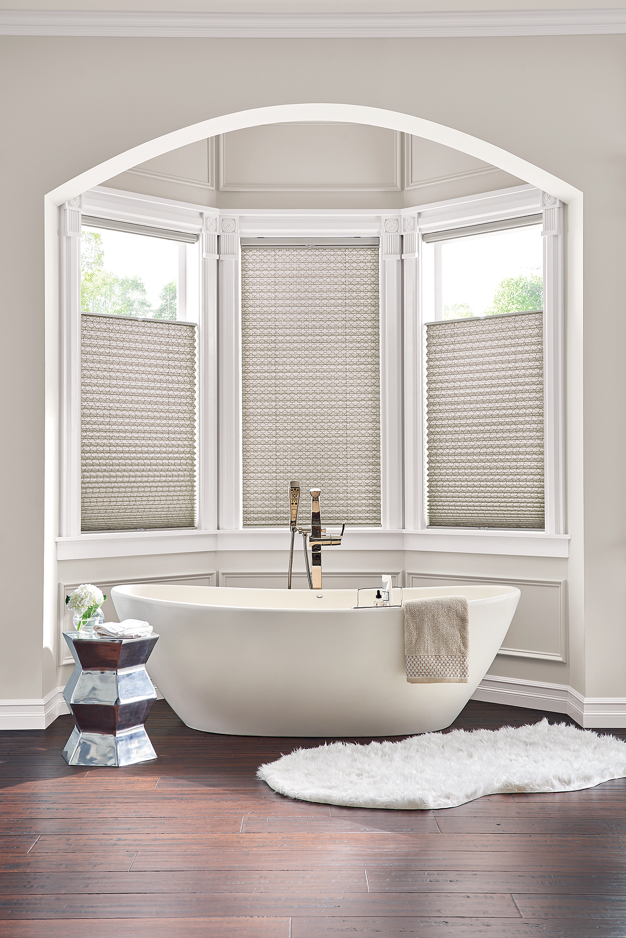 Elegant bathroom with a freestanding white tub positioned in front of bay windows, featuring light-filtering cellular shades for privacy and style.