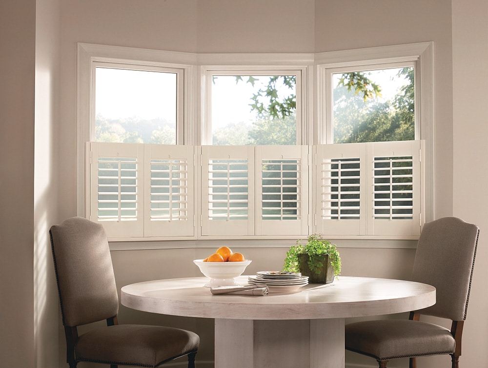 Cozy dining nook with a round table and two chairs in front of bay windows fitted with white café-style interior shutters.