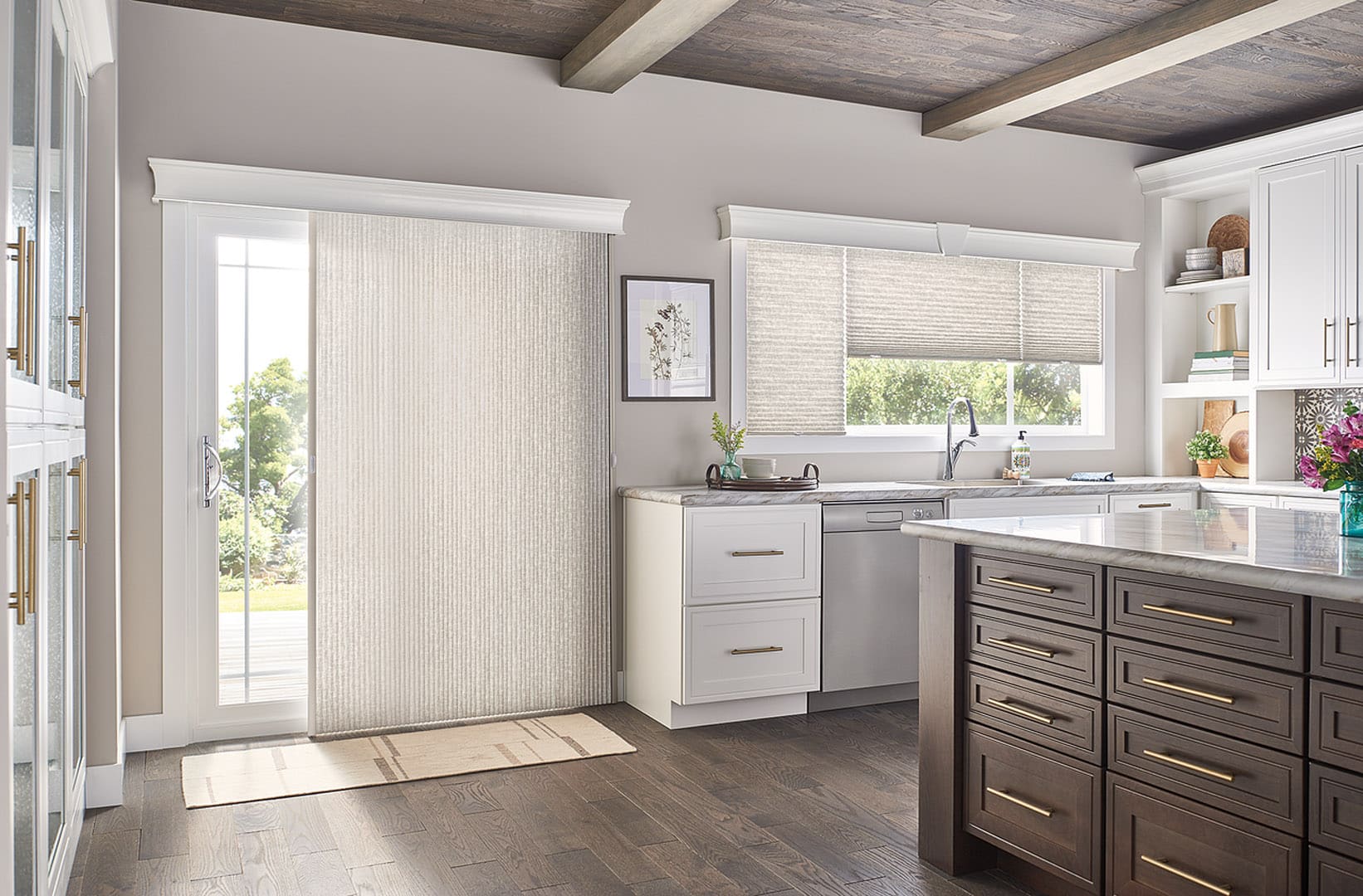 Modern kitchen with sliding glass door and window covered by light filtering cellular shades, featuring white cabinets, dark wood island with brass hardware, marble countertops, and wood beam ceiling.