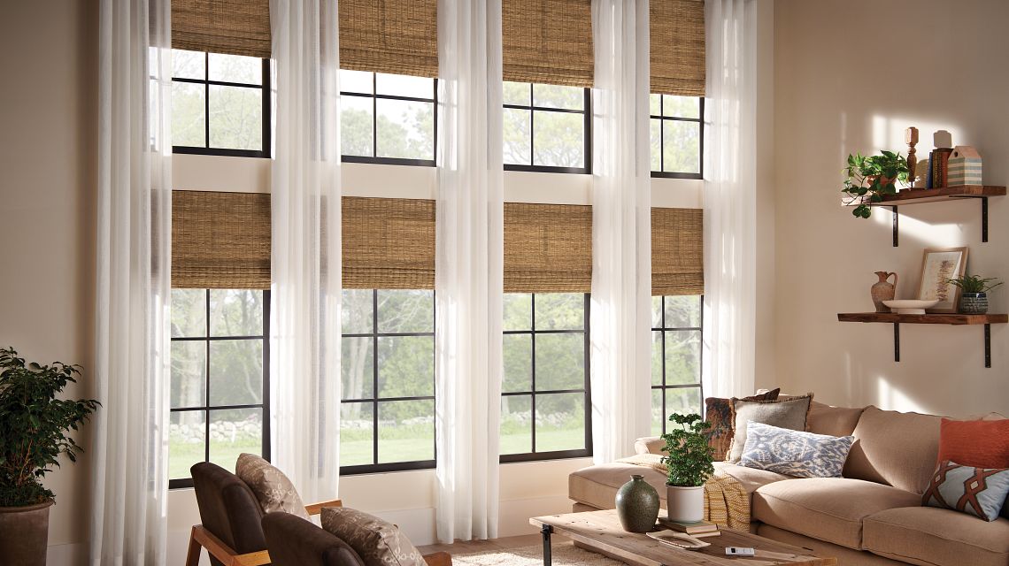 Light filled living room with tall black framed windows, sheer white curtains, and woven wood shades partially lowered.